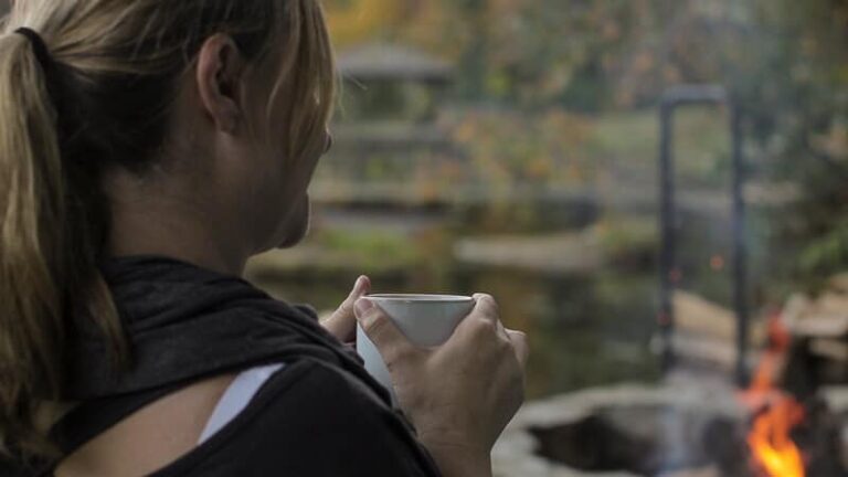 Woman holding a warm mug beside a fire pit near a lake